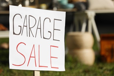 Photo of Sign Garage sale written on cardboard near tables with different stuff outdoors, closeup. Space for text