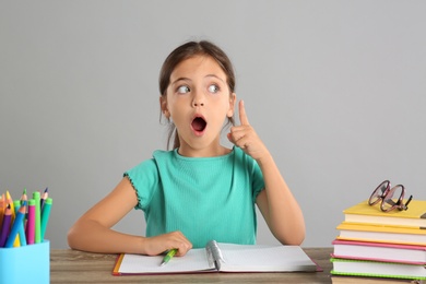 Emotional little girl doing homework at table on grey background