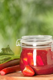 Jar of tasty rhubarb jam, fresh stems and strawberry on table against blurred background. Space for text