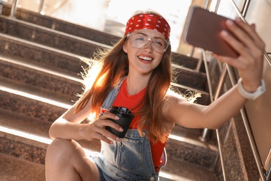 Photo of Beautiful young woman with coffee sitting on stairs and taking selfie outdoors