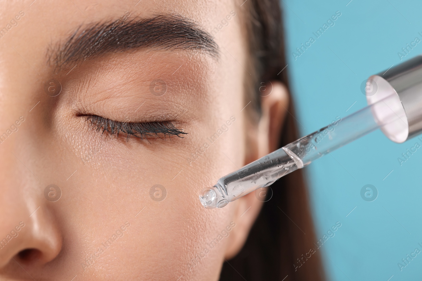 Photo of Young woman applying serum onto her face on light blue background, closeup