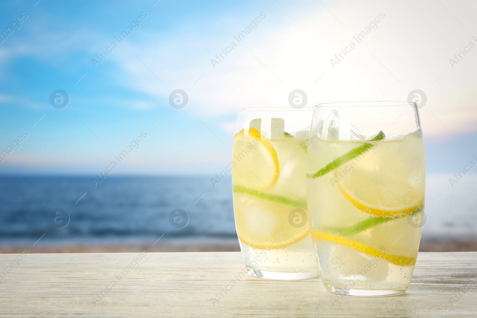 Image of Glasses of refreshing lemonade on wooden table near sea, space for text