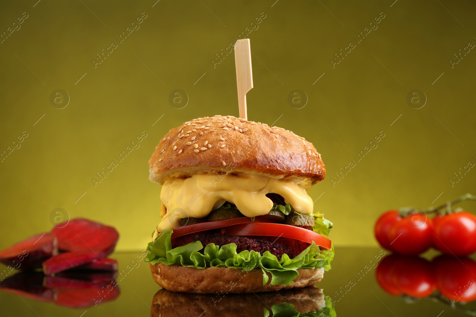 Photo of Delicious vegetarian burger, beetroot and tomatoes on mirror surface against olive background