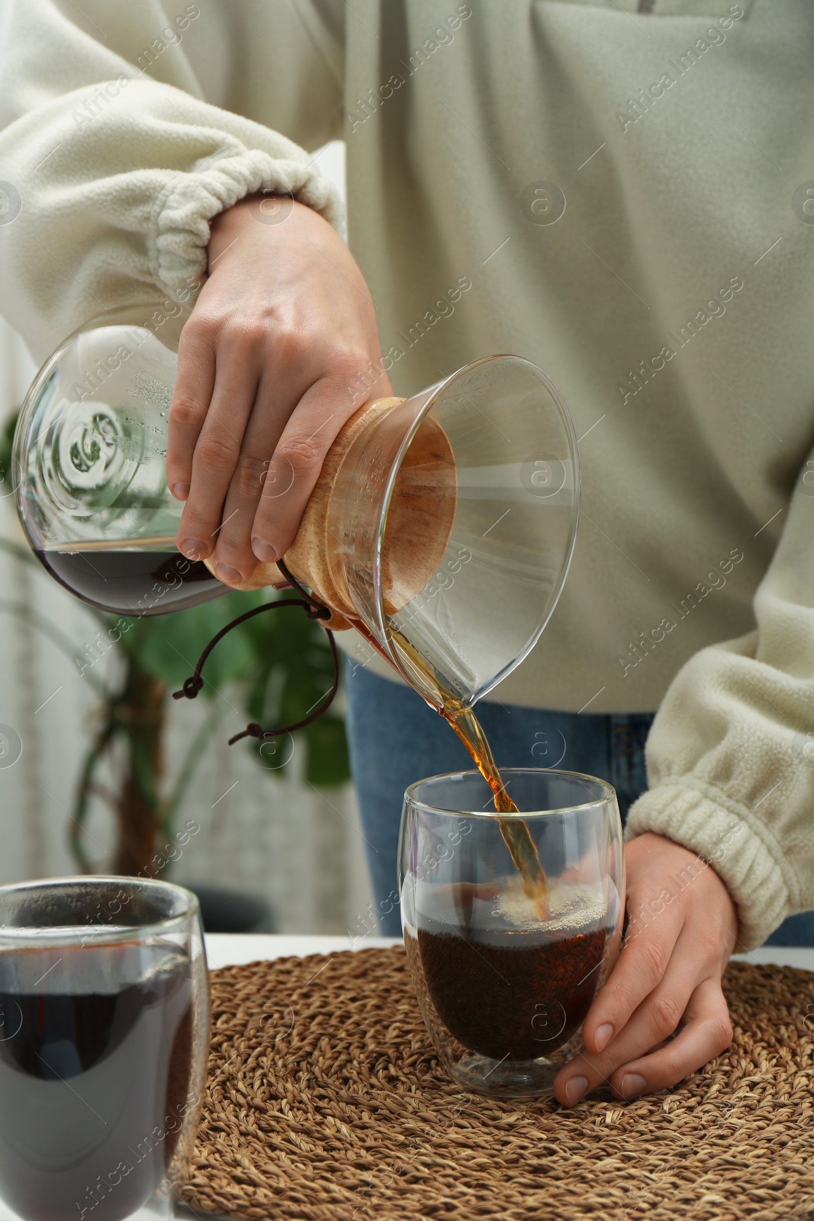 Photo of Woman pouring drip coffee from chemex coffeemaker into glass at table, closeup