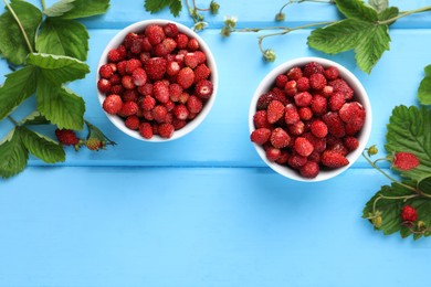 Fresh wild strawberries in bowls, flowers and leaves on light blue wooden table, flat lay. Space for text