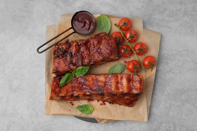 Photo of Tasty roasted pork ribs served with sauce, basil and tomatoes on grey table, top view