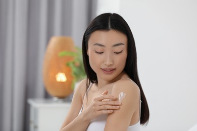 Photo of Beautiful young Asian woman applying body cream on shoulder in room