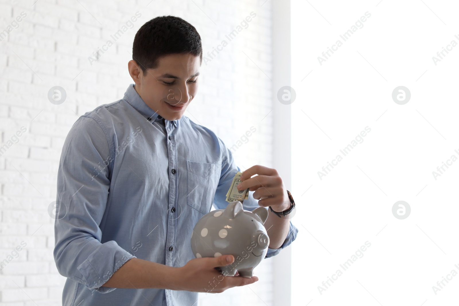 Photo of Young businessman putting money into piggy bank near brick wall. Space for text