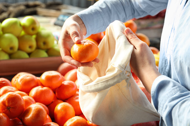 Photo of Woman putting tangerine into cotton eco bag at wholesale market, closeup. Life without plastic