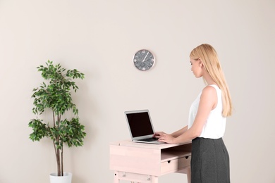 Photo of Young woman using laptop at stand up workplace against light wall