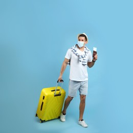 Male tourist in protective mask holding passport with ticket and suitcase on turquoise background
