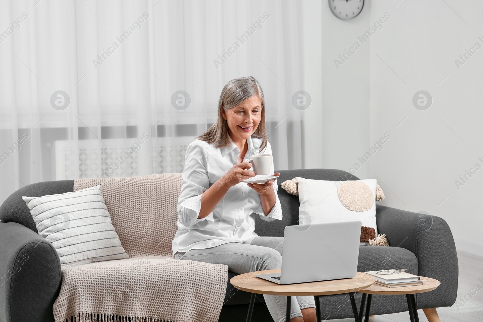 Photo of Beautiful senior woman with cup of drink near laptop at home