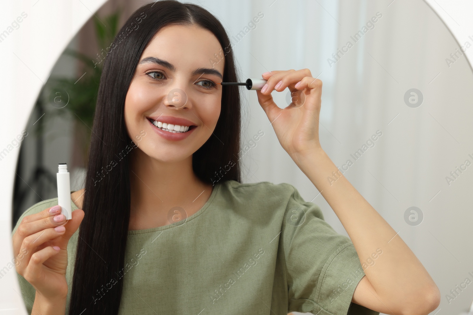 Photo of Beautiful woman applying serum onto her eyelashes in room, reflection in mirror. Cosmetic product