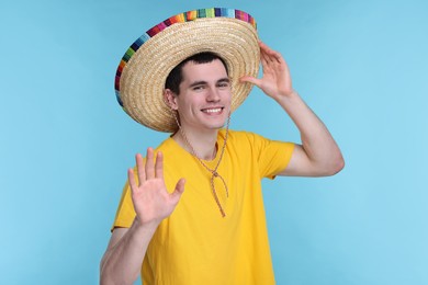 Photo of Young man in Mexican sombrero hat waving hello on light blue background