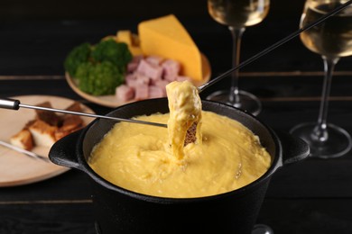 Photo of Dipping pieces of ham and bread into fondue pot with melted cheese on black wooden table, closeup