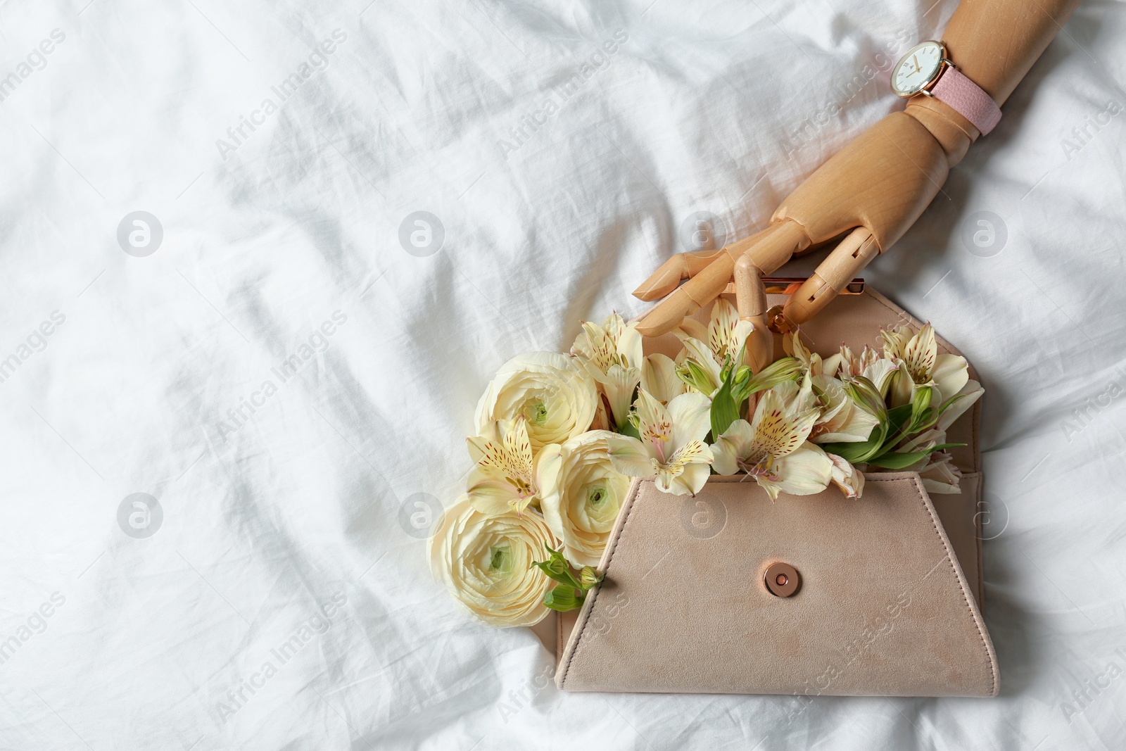 Photo of Stylish clutch with spring flowers and wooden mannequin hand on crumpled sheet, flat lay. Space for text