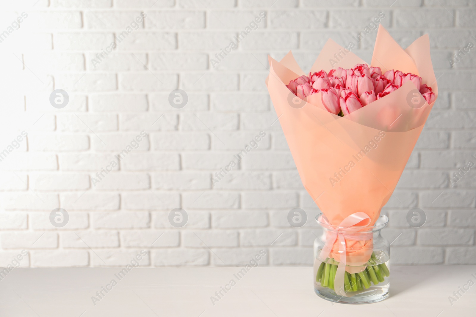 Photo of Bouquet of beautiful pink tulips in vase on white table near brick wall, space for text