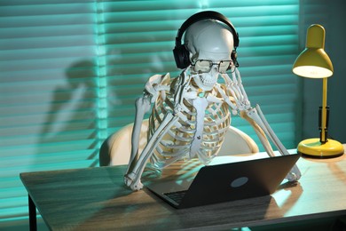 Waiting concept. Human skeleton sitting at wooden table with laptop and headphones indoors