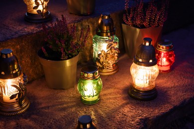 Photo of Different grave lanterns with burning candles on stone surface at night