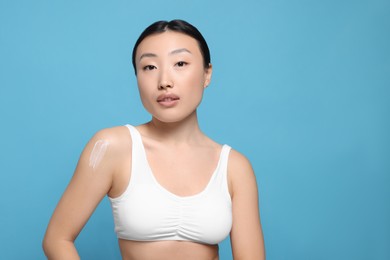 Photo of Beautiful young Asian woman with smear of body cream on shoulder against light blue background, space for text