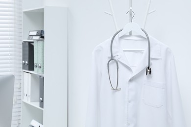 White doctor's gown and stethoscope hanging on rack in clinic, closeup. Space for text