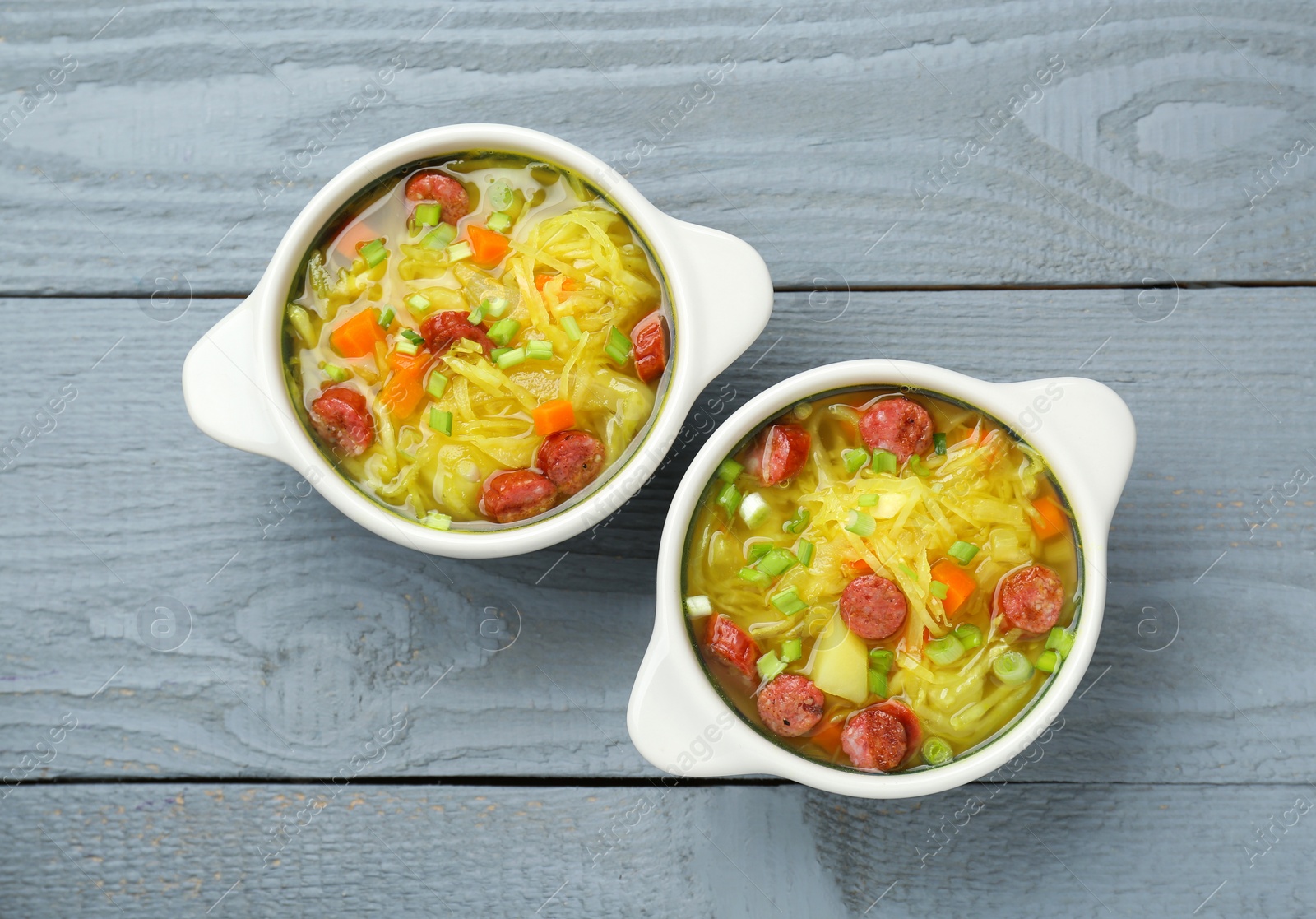 Photo of Delicious sauerkraut soup with smoked sausages and green onion on grey wooden table, flat lay