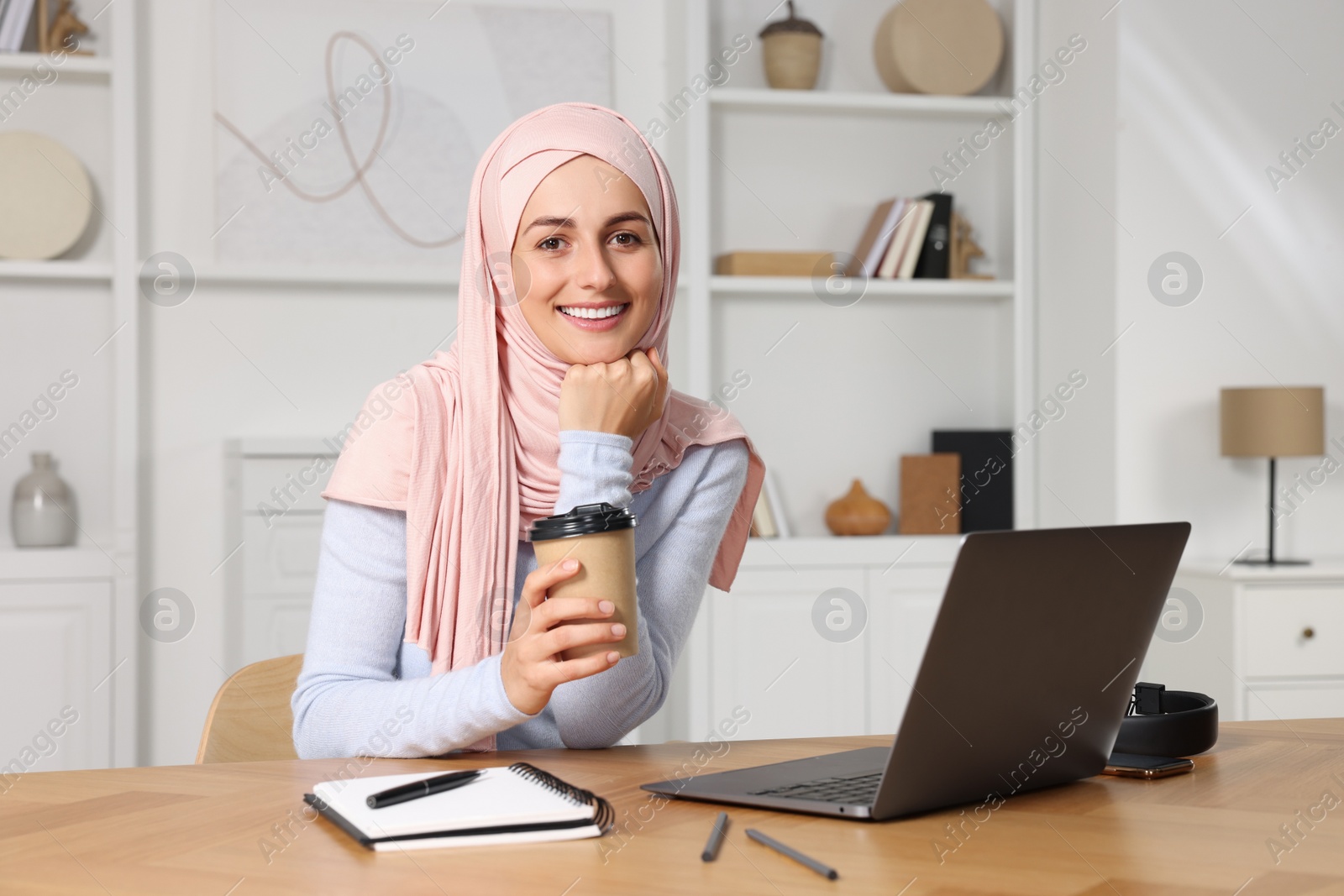 Photo of Muslim woman with cup of coffee using laptop at wooden table in room