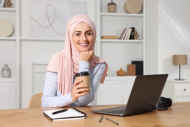 Muslim woman with cup of coffee using laptop at wooden table in room