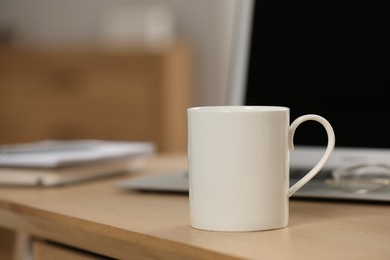 White ceramic mug and laptop on wooden table at workplace. Space for text