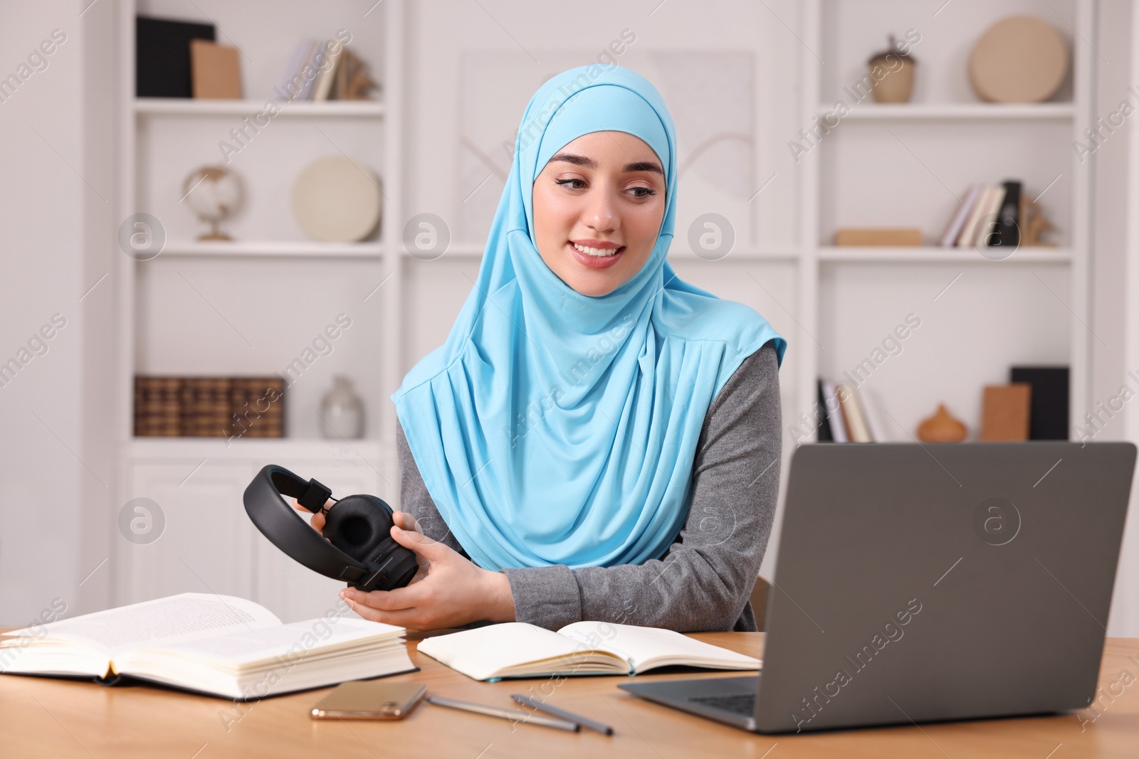 Photo of Muslim woman in hijab with headphones using laptop at wooden table indoors