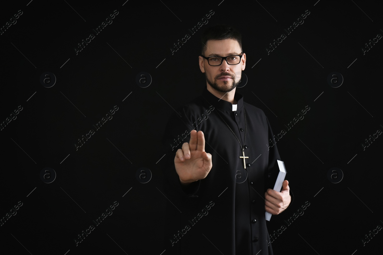 Photo of Priest with Bible making blessing gesture on dark background, space for text
