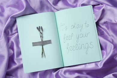 Photo of Phrase It`s Okay to Feel Your Feelings and dry lavender attached with adhesive tape in notebook on violet fabric, top view