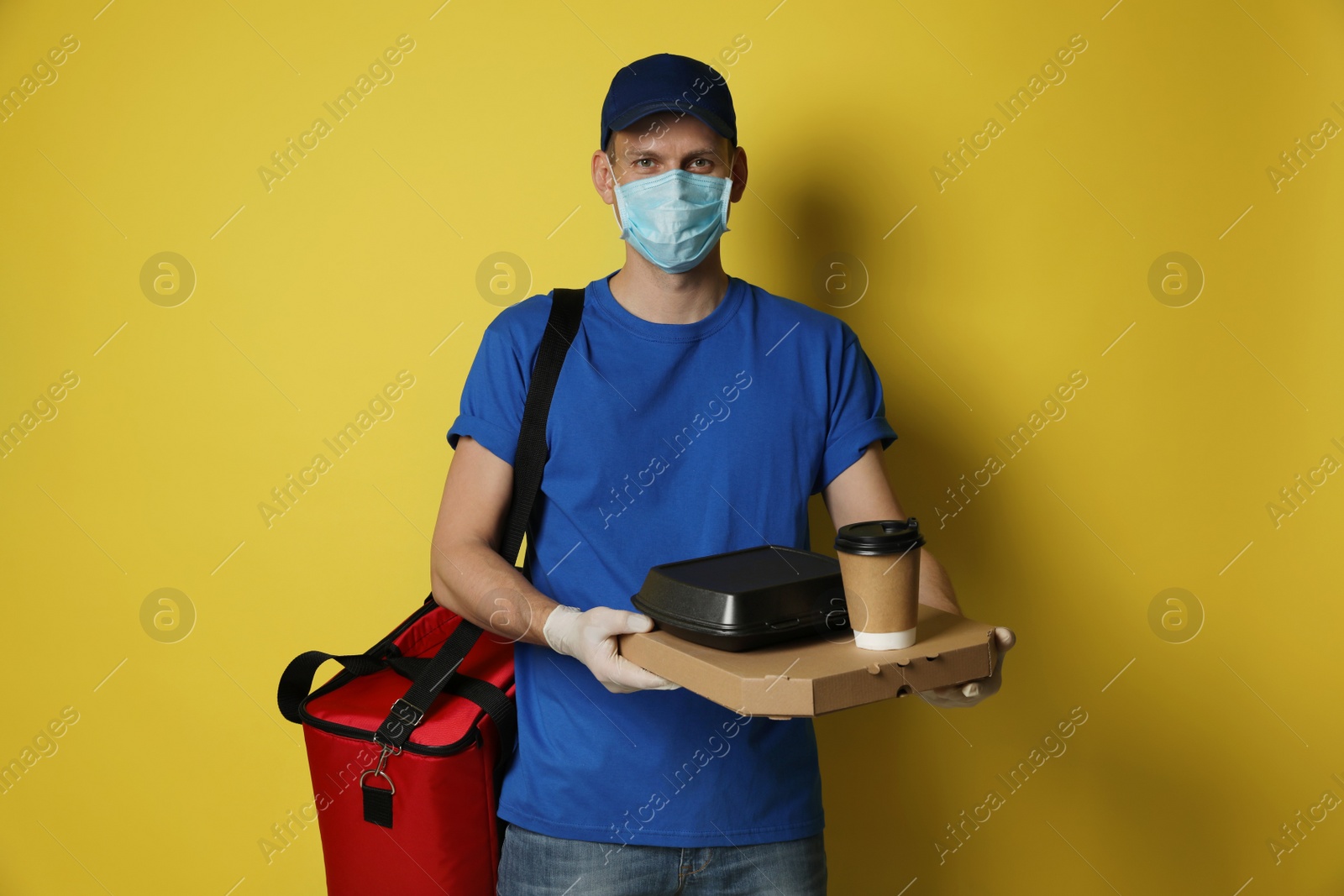 Photo of Courier in protective mask and gloves holding order on yellow background. Food delivery service during coronavirus quarantine