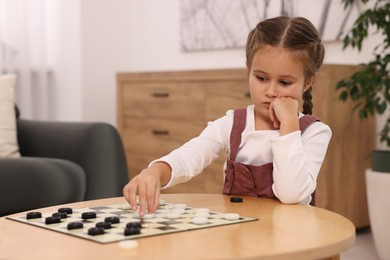 Thoughtful girl playing checkers at wooden table in room