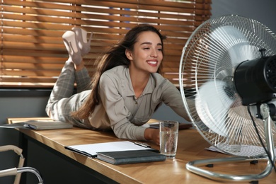 Photo of Woman enjoying air flow from fan while lying on table at workplace. Summer heat