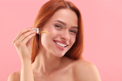 Beautiful young woman applying cosmetic serum onto her face on pink background