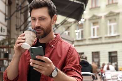 Handsome man with cup of coffee using smartphone outdoors, space for text