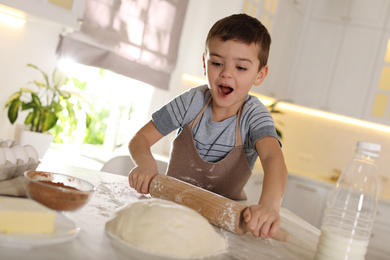 Photo of Emotional little boy rolling dough at table in kitchen. Cooking pastry