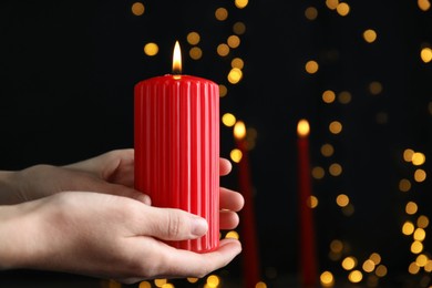 Photo of Woman holding burning red candle against blurred Christmas lights, closeup. Space for text