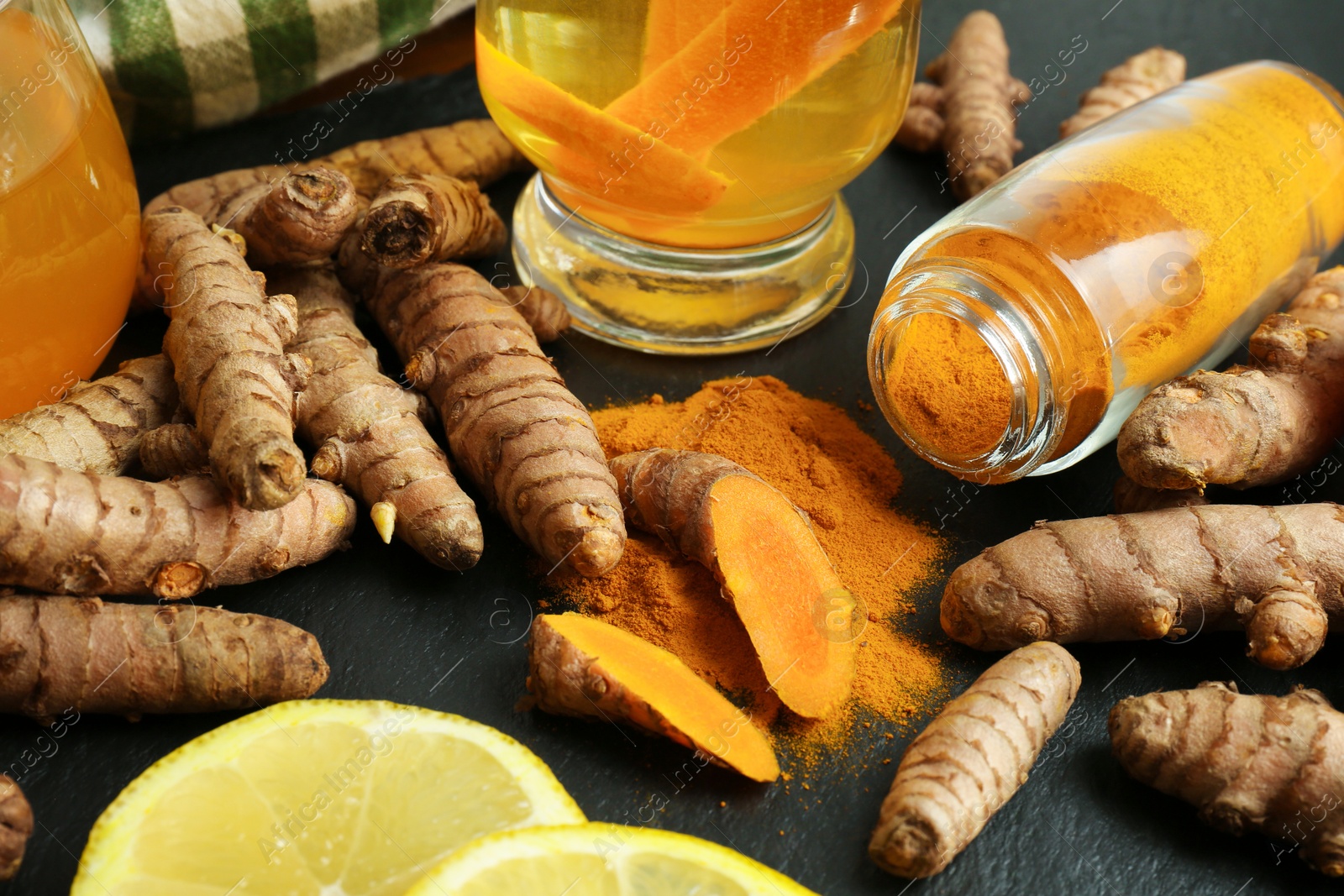 Photo of Turmeric roots, powder, sliced lemon and cup of tasty tea on black textured table, closeup