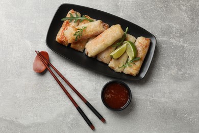 Photo of Tasty fried spring rolls, arugula, lime and sauce served on grey textured table, flat lay