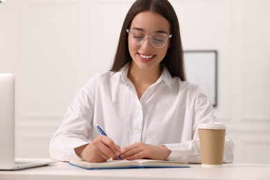 Photo of Young woman writing in notebook at white table indoors