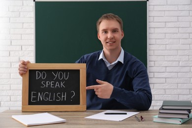 Teacher holding small chalkboard with inscription Do You Speak English? at table in classroom