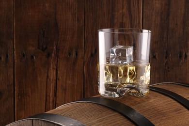 Whiskey with ice cubes in glass on barrel against wooden background, space for text