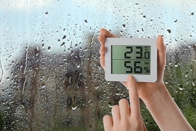 Image of Woman holding digital hygrometer with thermometer near window on rainy day, closeup. Space for text