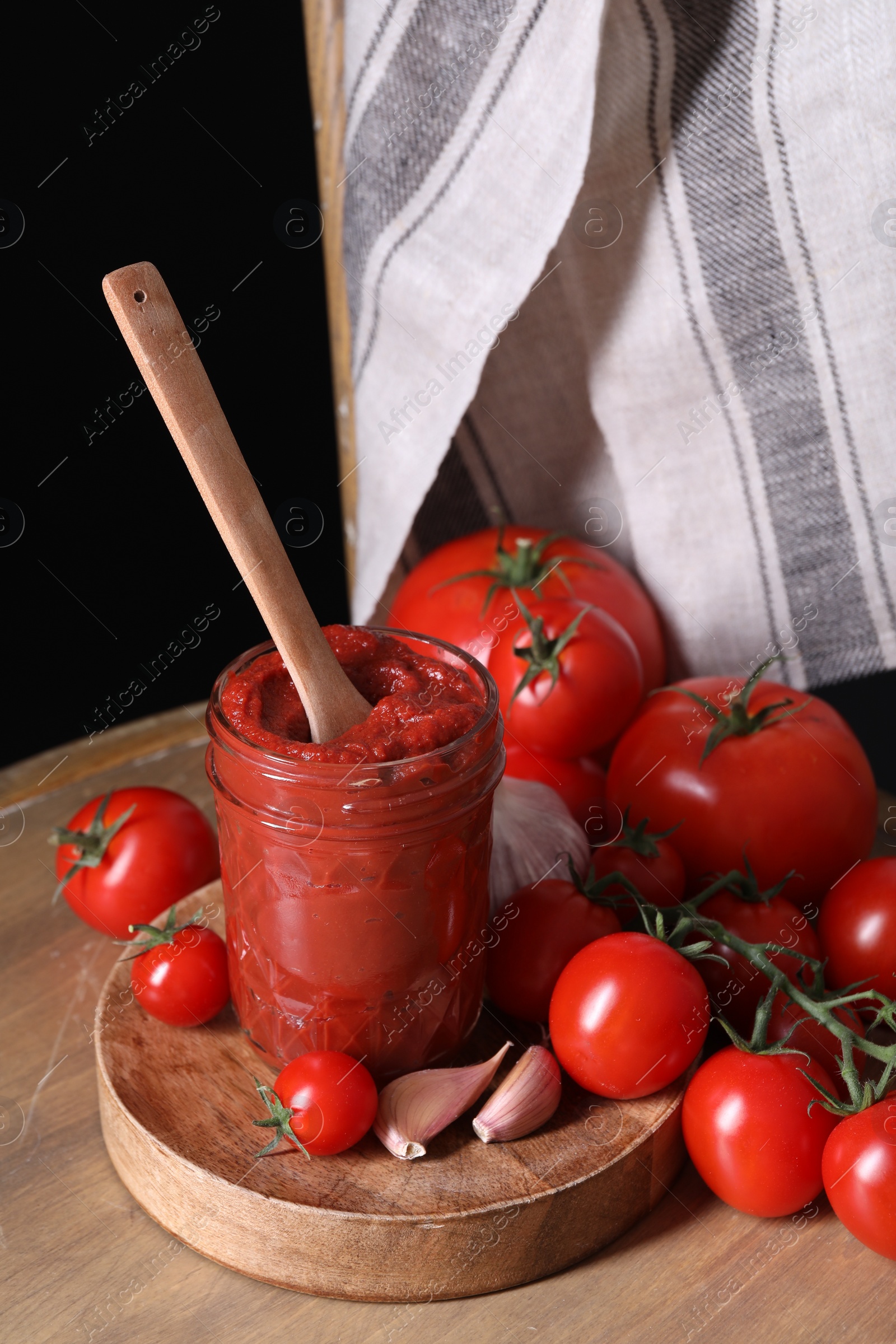 Photo of Jar of tasty tomato paste with spoon and ingredients on wooden table