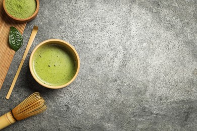 Photo of Cup of fresh matcha tea, bamboo whisk, spoon and green powder on grey table, flat lay. Space for text