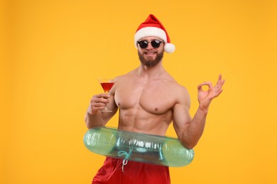 Photo of Muscular young man in Santa hat with inflatable ring and cocktail showing ok gesture on orange background