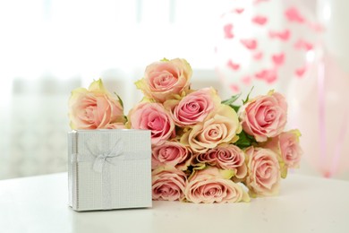 Photo of Beautiful bouquet of roses and gift on white table in room. Happy birthday greetings