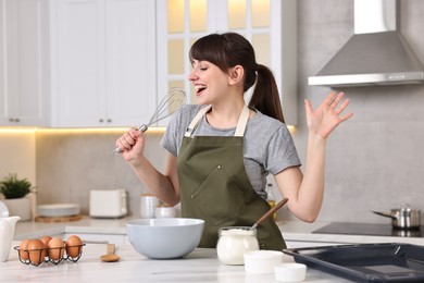 Happy young housewife with whisk having fun while cooking at white marble table in kitchen